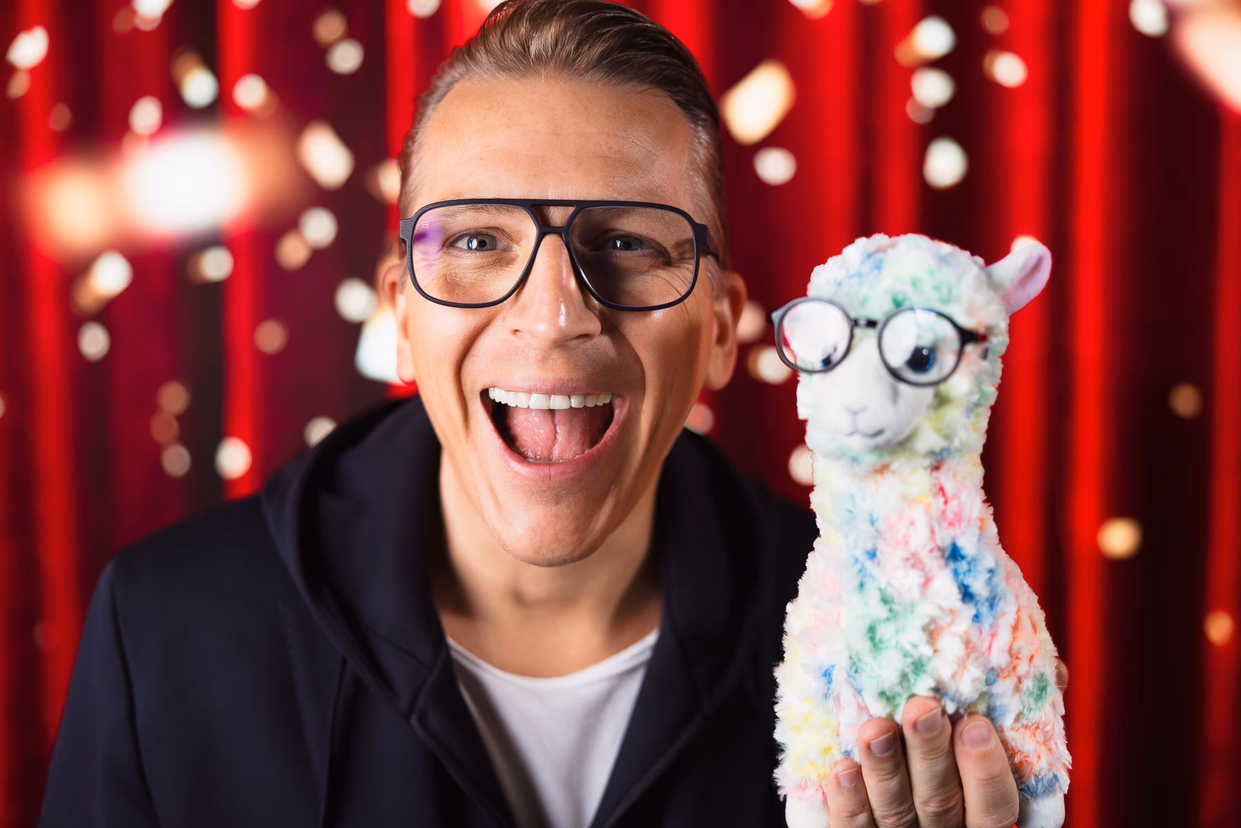 Comedian mit Lama-Plüschtier vor rotem Bühnenvorhang, breites Lachen